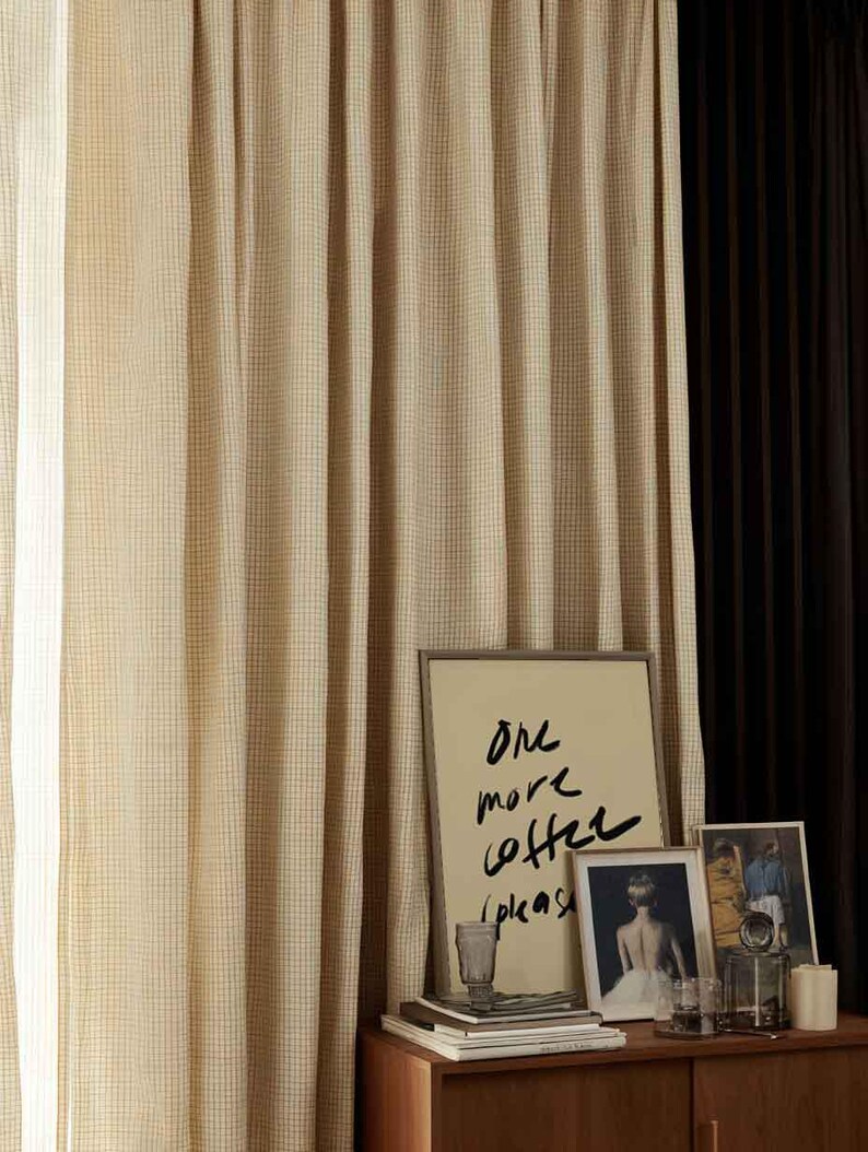 Puede incluir: Cortinas beige y marr&oacute;n a cuadros cuelgan junto a una cortina marr&oacute;n oscuro. Una mesa de madera contiene arte enmarcado, incluida una impresi&oacute;n con el texto "One more coffee please". Otros art&iacute;culos incluyen un vaso, libros y una vela.