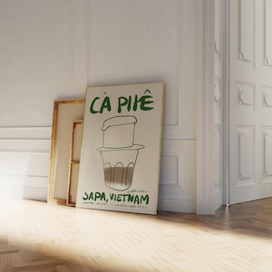 May include: A framed poster with the words "C&agrave; Ph&ecirc;" in green, depicting a Vietnamese coffee filter and cup. The text "Sapa, Vietnam" is below the illustration, with additional text about the coffee. The poster leans against a wall.