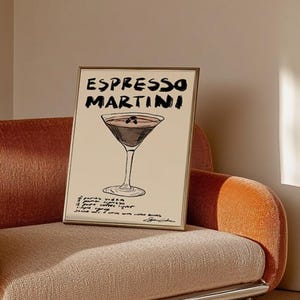 May include: A framed print with an Espresso Martini design. The words "ESPRESSO MARTINI" are written in black above a cocktail illustration. The recipe is below the image. The frame is gold. The print is displayed on a peach-colored sofa.