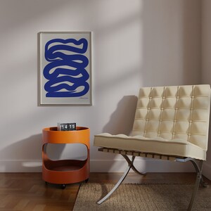 May include: A white chair with a tufted back and a chrome base sits in a room with a white wall and a framed print of a blue abstract design. A small orange side table with a clock on top is in the foreground.