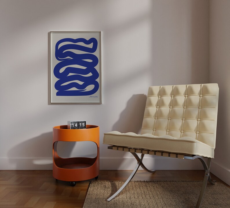 May include: A white chair with a tufted back and a chrome base sits in a room with a white wall and a framed print of a blue abstract design. A small orange side table with a clock on top is in the foreground.