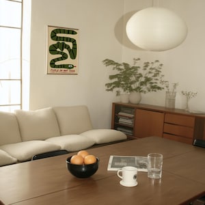 May include: A dining room with a dark wood table set with a black bowl of oranges, a newspaper, a mug, and a glass. A cream sofa and wooden cabinet are in the background. A poster with a green snake and the text "PLEASE DO NOT FEED" hangs on the wall.