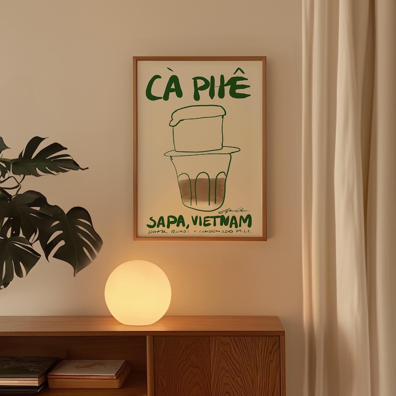 May include: A framed poster with the words "C&agrave; Ph&ecirc;" in green, depicting a Vietnamese coffee filter and the text "Sapa, Vietnam." A round, glowing lamp sits on a wooden cabinet below the poster.