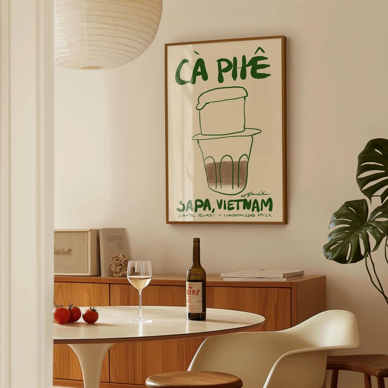 May include: A framed poster with green text and a coffee illustration, reading "CA PHE" and "SAPA, VIETNAM." A bottle of wine, a glass of wine, and three tomatoes sit on a white table. A wooden cabinet and a plant are also visible.