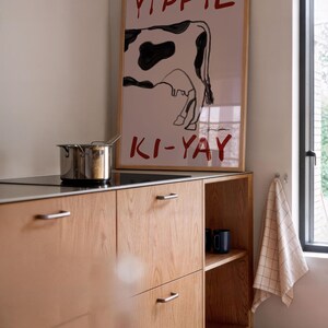 Op de afbeelding: Een keukeninterieur met een houten kast en een ingelijste poster met een koe-illustratie en de rode woorden "YIPPIE KI-YAY". Een roestvrijstalen pot staat op de kookplaat. De vloer is betegeld.