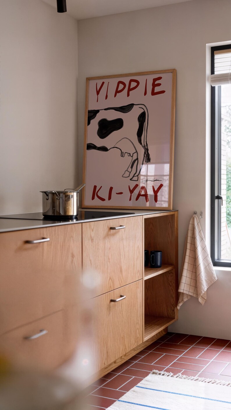 Op de afbeelding: Een keukeninterieur met een houten kast en een ingelijste poster met een koe-illustratie en de rode woorden "YIPPIE KI-YAY". Een roestvrijstalen pot staat op de kookplaat. De vloer is betegeld.