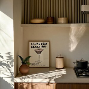 May include: A kitchen interior with a framed "Espresso Martini" print, a small brown vase with greenery, and a cylindrical container. A pot sits on a hob. The colour scheme is neutral, with natural light creating a warm atmosphere.