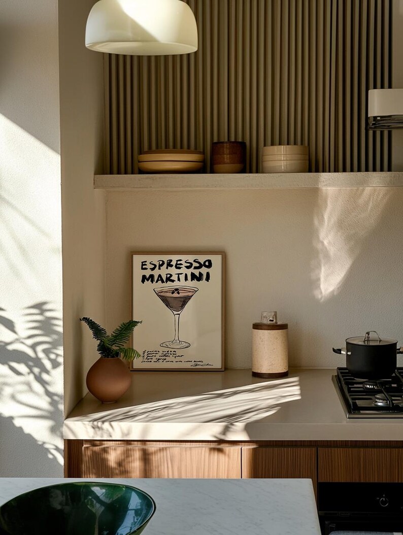 May include: A kitchen interior with a framed "Espresso Martini" print, a small brown vase with greenery, and a cylindrical container. A pot sits on a hob. The colour scheme is neutral, with natural light creating a warm atmosphere.