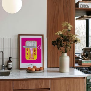 May include: A framed rectangular art print featuring a vibrant magenta background with a yellow sardine tin graphic. The illustration depicts a silver fish inside the tin with the text PORTUGUESE SARDINES printed vertically. The print is displayed on a kitchen counter next to a beige ceramic vase containing dried flowers and a small bowl of brown eggs.