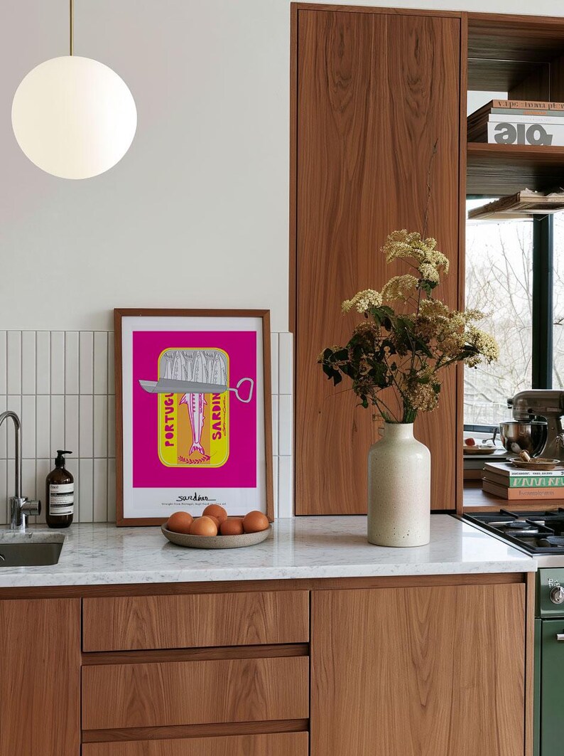 May include: A framed rectangular art print featuring a vibrant magenta background with a yellow sardine tin graphic. The illustration depicts a silver fish inside the tin with the text PORTUGUESE SARDINES printed vertically. The print is displayed on a kitchen counter next to a beige ceramic vase containing dried flowers and a small bowl of brown eggs.