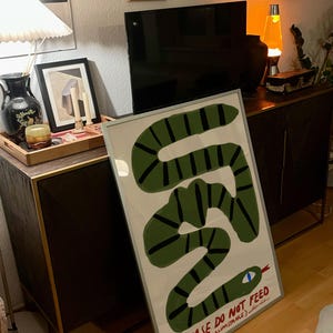 May include: A framed art print depicting a green snake with black stripes on a white backdrop. The snake has a blue eye and a red tongue. The text "PLEASE DO NOT FEED (UNDER ANY CIRCUMSTANCE)" is at the bottom. The artwork is displayed in a room with other decor.