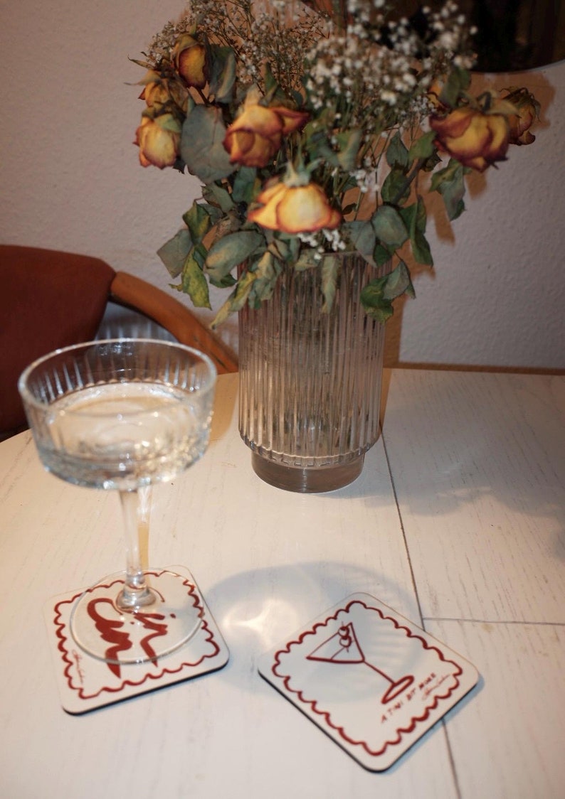 May include: A clear coupe glass with liquid sits on a white coaster with a red border and a cocktail illustration. A second coaster with a similar design is next to it. A vase of dried flowers is in the background.