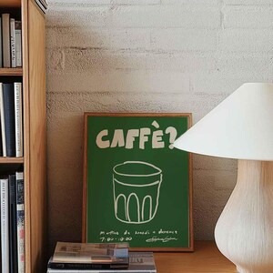 Op de afbeelding: Een groene poster met het woord "CAFF&Egrave;?" in het wit en een tekening van een koffiekopje. De poster staat op een houten bureau met boeken en een lamp. Links staat een houten boekenkast.