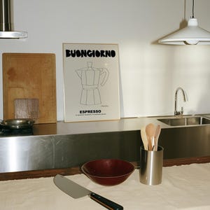 May include: A framed wall print featuring a black line drawing of a moka pot with the text BUONGIORNO and ESPRESSO, positioned above a kitchen counter with a dark red bowl, a metal utensil holder with wooden spoons, and a large chef's knife.