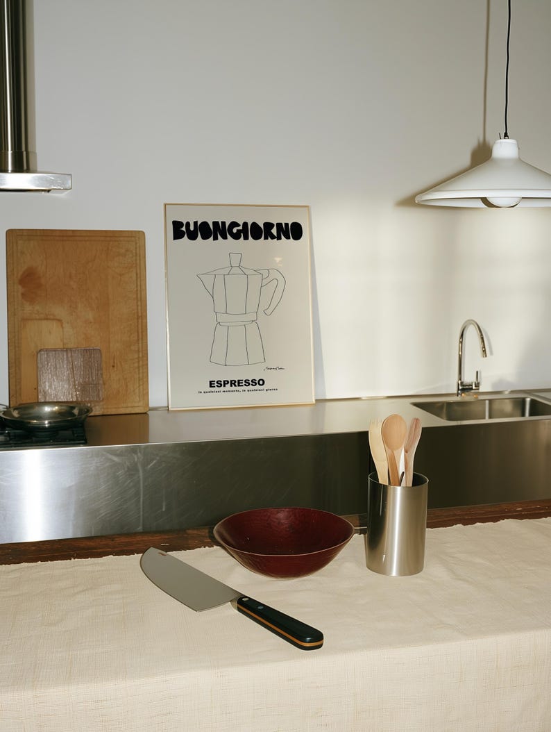 May include: A framed wall print featuring a black line drawing of a moka pot with the text BUONGIORNO and ESPRESSO, positioned above a kitchen counter with a dark red bowl, a metal utensil holder with wooden spoons, and a large chef's knife.