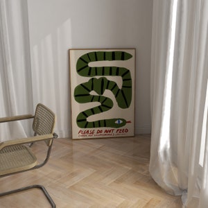 May include: A framed art print depicting a green snake with black stripes on a cream backdrop. The artwork includes the text "PLEASE DO NOT FEED (UNDER ANY CIRCUMSTANCE)". The snake has a blue eye and a red tongue.