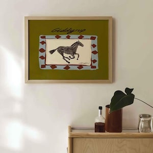 Puede incluir: Una impresi&oacute;n art&iacute;stica enmarcada con una ilustraci&oacute;n de un caballo corriendo. La obra de arte tiene un fondo verde con un dise&ntilde;o rectangular en azul y rojo. Las palabras "Giddy-up" est&aacute;n escritas encima del caballo.