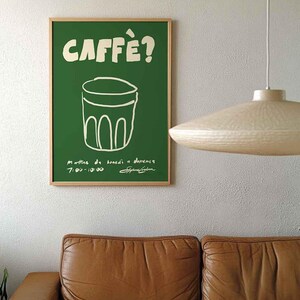 Op de afbeelding: Een groene poster met het woord "CAFF&Egrave;?" in het wit, boven een witte lijntekening van een glas. De poster zit in een houten lijst en hangt aan een witte muur. Daaronder staat een bruine leren bank.