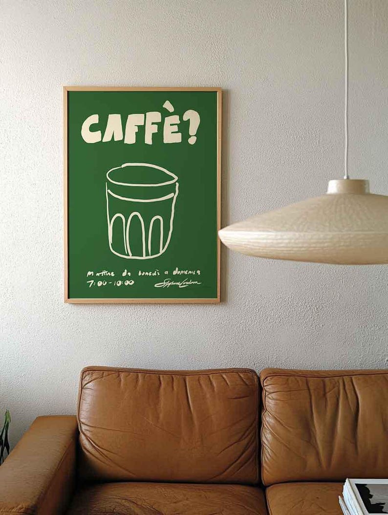 Op de afbeelding: Een groene poster met het woord "CAFF&Egrave;?" in het wit, boven een witte lijntekening van een glas. De poster zit in een houten lijst en hangt aan een witte muur. Daaronder staat een bruine leren bank.