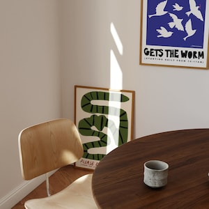 May include: A room corner with framed art prints and a wooden chair. One print shows a green and black snake design, another displays white birds on a blue background with the text "GETS THE WORM". A small grey ceramic cup sits on a round wooden table.