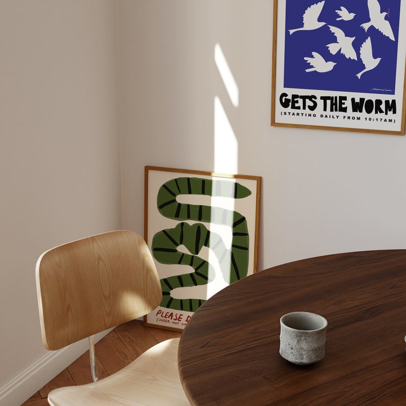 May include: A room corner with framed art prints and a wooden chair. One print shows a green and black snake design, another displays white birds on a blue background with the text "GETS THE WORM". A small grey ceramic cup sits on a round wooden table.