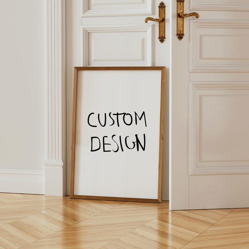 以下が含まれることがあります： 「CUSTOM DESIGN」の文字が黒字で書かれたフレーム入りのプリント。フレームは温かみのある茶色で、プリントは白いドアと壁に立てかけられています。床は木製です。