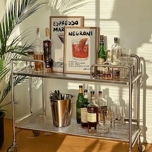 May include: A chrome bar cart with two tiers, stocked with bottles of spirits, cocktail glasses, and an ice bucket. Two framed prints, one reading "ESPRESSO MARTINI" and the other "NEGRONI", are displayed on the top shelf.