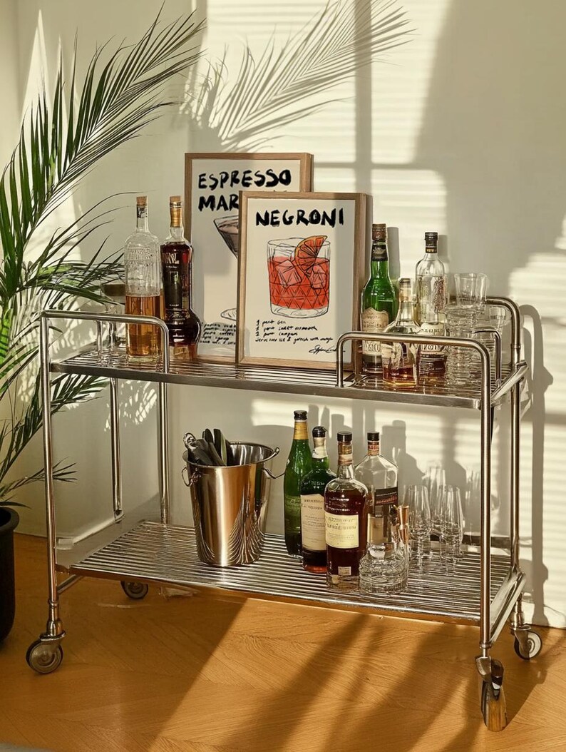 May include: A chrome bar cart with two tiers, stocked with bottles of spirits, cocktail glasses, and an ice bucket. Two framed prints, one reading "ESPRESSO MARTINI" and the other "NEGRONI", are displayed on the top shelf.
