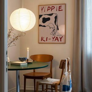 Op de afbeelding: Eetkamer met een ronde groene tafel, houten stoelen en een poster met een koe-illustratie en de tekst "YIPPIE KI-YAY". Een papieren lantaarn hangt aan het plafond. Een brandende kaars en een kom staan op de tafel.