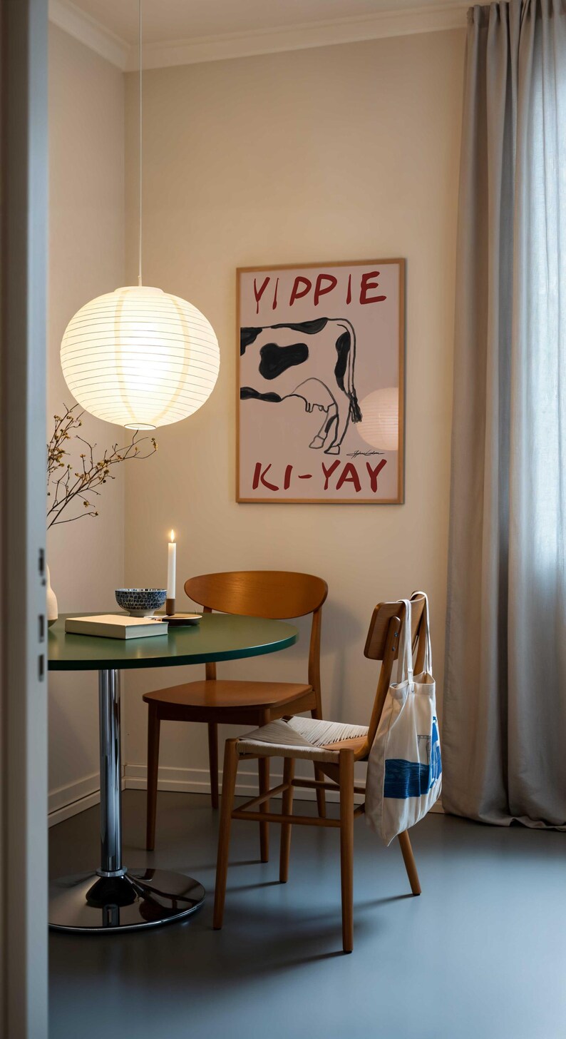 Op de afbeelding: Eetkamer met een ronde groene tafel, houten stoelen en een poster met een koe-illustratie en de tekst "YIPPIE KI-YAY". Een papieren lantaarn hangt aan het plafond. Een brandende kaars en een kom staan op de tafel.