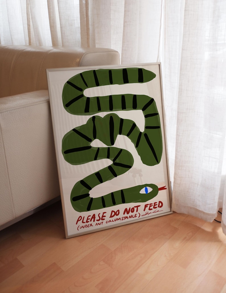 May include: A framed art print depicting a green snake with black stripes and a blue eye. The text "PLEASE DO NOT FEED (UNDER ANY CIRCUMSTANCE)" is written below the snake. The artwork is in a white frame, suitable for home decor.