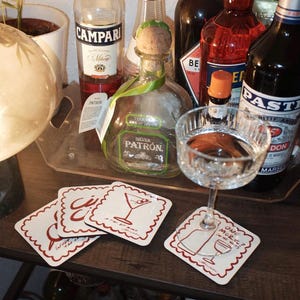 May include: A bar setup featuring various liquor bottles, including Campari and Patron. Several white coasters with red illustrations of cocktails and text are visible, along with a stemmed glass. A small plant and a lamp are also in the scene.