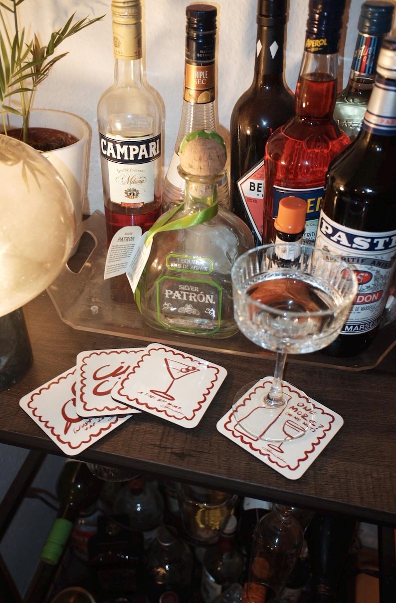 May include: A bar setup featuring various liquor bottles, including Campari and Patron. Several white coasters with red illustrations of cocktails and text are visible, along with a stemmed glass. A small plant and a lamp are also in the scene.