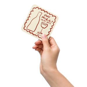May include: A square coaster with a scalloped edge, featuring a wine bottle and glass illustration in brown. The text "ONE MORE? (WINE NOT)" is written above the illustrations. The coaster is held by a hand against a white background.