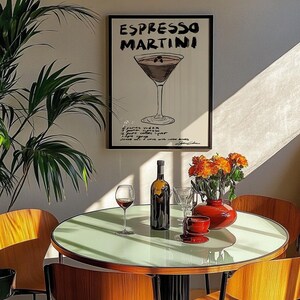 May include: A stylish dining area with a round table and wooden chairs. A framed poster reading "Espresso Martini" is on the wall. A bottle of wine, a glass of red wine, and a red vase with orange flowers are on the table.