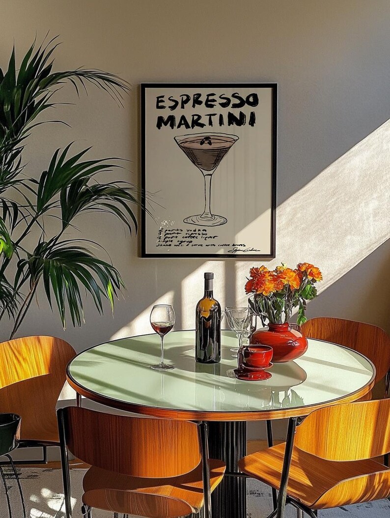 May include: A stylish dining area with a round table and wooden chairs. A framed poster reading "Espresso Martini" is on the wall. A bottle of wine, a glass of red wine, and a red vase with orange flowers are on the table.