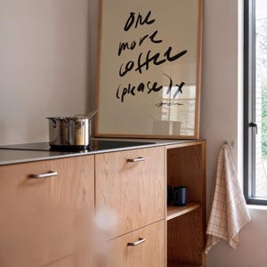 Puede incluir: Una cocina con gabinetes de madera clara y un cartel enmarcado con la frase "One more coffee (please)". Una olla de acero inoxidable est&aacute; en la estufa. Dos tazas azul oscuro est&aacute;n en un estante.