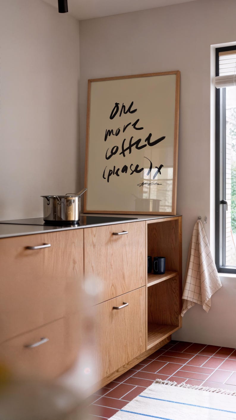 Puede incluir: Una cocina con gabinetes de madera clara y un cartel enmarcado con la frase "One more coffee (please)". Una olla de acero inoxidable est&aacute; en la estufa. Dos tazas azul oscuro est&aacute;n en un estante.