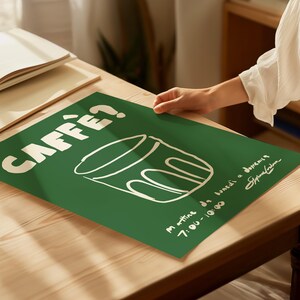 Op de afbeelding: Groene poster met het woord "CAFF&Egrave;?" in het wit, samen met een lijntekening van een koffiekopje. De poster bevat ook tekst in het Italiaans en een handtekening. De poster ligt op een houten tafel.