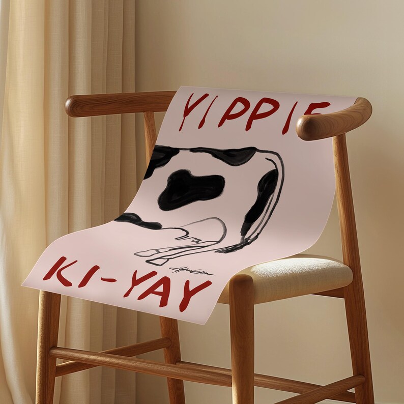 Op de afbeelding: Een roze poster met een zwart-witte koe-illustratie en de woorden "YIPPIE" en "KI-YAY" in rode letters. De poster is over een houten stoel met een lichtgekleurd kussen gedrapeerd.