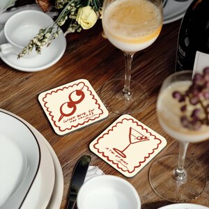 May include: Two square coasters with scalloped edges sit on a wooden table. One features a red olive design, the other a martini glass. Text on the coasters reads "(Just here for the snacks?)" and "A Tini Bit More."