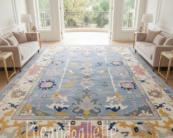May include: A large, rectangular area rug with a light blue central field and a detailed border. The rug features a floral and geometric pattern in shades of pink, yellow, and gray. The rug is placed in a bright, open room with two sofas.