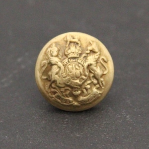 May include: A round, antique-style button with a detailed embossed design. The button features a central crest with a crown, flanked by two heraldic lions. The button is a light gold color.