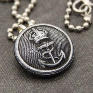 May include: A silver chain necklace with a black button pendant featuring a silver crown and anchor design.