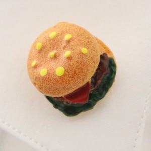 May include: A novelty cufflink shaped like a miniature hamburger. The burger has a sesame seed bun, a patty, lettuce, and a slice of tomato. The cufflink is resting on a white shirt cuff, with a striped tie visible in the background.