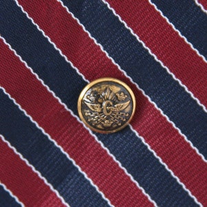 May include: A gold tie tack with a crest featuring a letter "G" surrounded by wings and a crown. The tie tack is pinned to a red and navy blue striped tie.