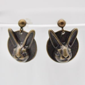 May include: A pair of bronze-colored stud earrings. Each earring features a detailed rabbit head design within a circular frame. The earrings have a post and ball closure. The rabbit's ears are upright, and the face is detailed.