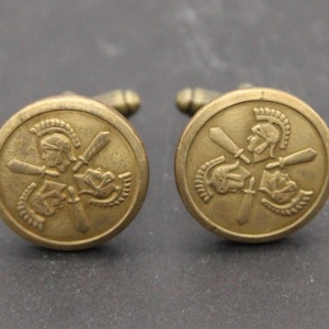 May include: A pair of antique brass cufflinks. Each cufflink features a circular design with a detailed relief of a Roman soldier in a helmet, holding a sword. The cufflinks have a classic, vintage aesthetic.