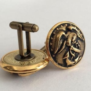 May include: A pair of gold-toned cufflinks with a detailed design. Each cufflink features a circular face with an embossed eagle and anchor motif. The cufflinks have a classic T-bar closure.