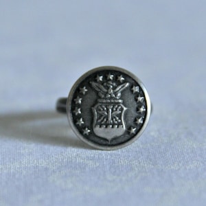 Puede incluir: Un anillo de plata con un emblema de la Fuerza Aérea de los Estados Unidos. El emblema presenta un escudo con un águila alada y una estrella.